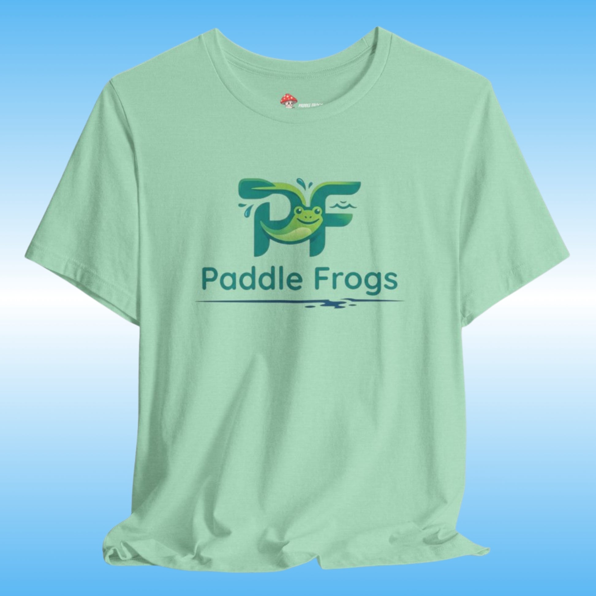 A light mint green t-shirt featuring a green and blue 'Paddle Frogs' tadpole printed on the chest, displayed against a blue gradient background.