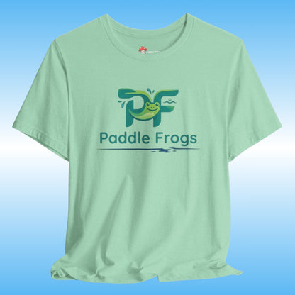 A light mint green t-shirt featuring a green and blue 'Paddle Frogs' tadpole printed on the chest, displayed against a blue gradient background.