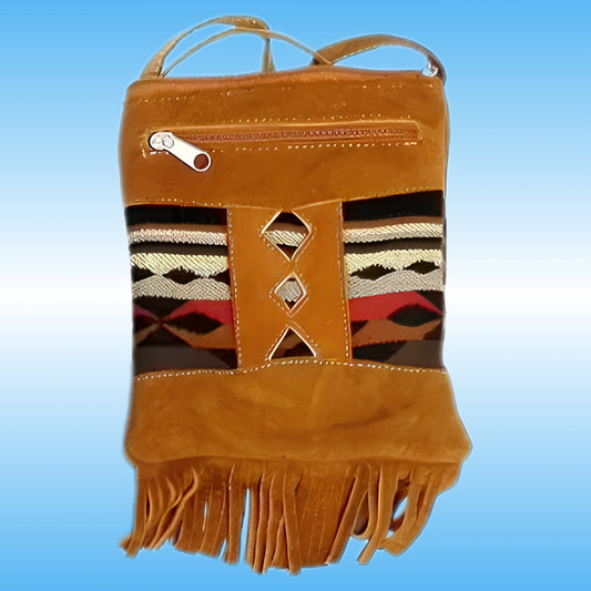 A tan leather crossbody bag with a Southwestern-style woven pattern panel on the front, featuring geometric stripes in black, white, red, and brown, and a fringed bottom edge.