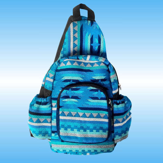 A blue patterned sling backpack with geometric Southwestern-style woven designs in shades of blue, teal, and white, featuring black straps and zippers, set against a light blue gradient background.