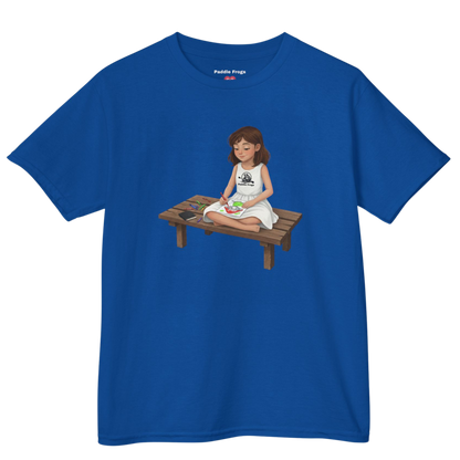 Blue t-shirt with a graphic of Willow, Paddle Frogs character sitting on a wooden bench coloring Bailey on a transparent background