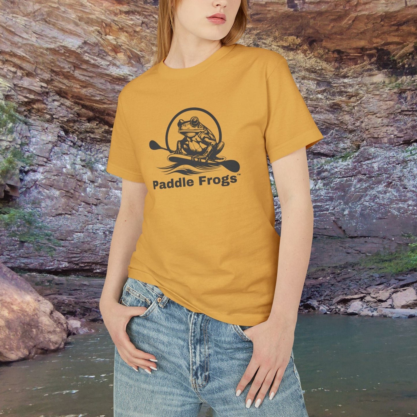 Woman wearing a yellow 'Paddle Frogs' t-shirt standing in front of a natural rock formation. Earthy Tee collection Faded mustard Yellow