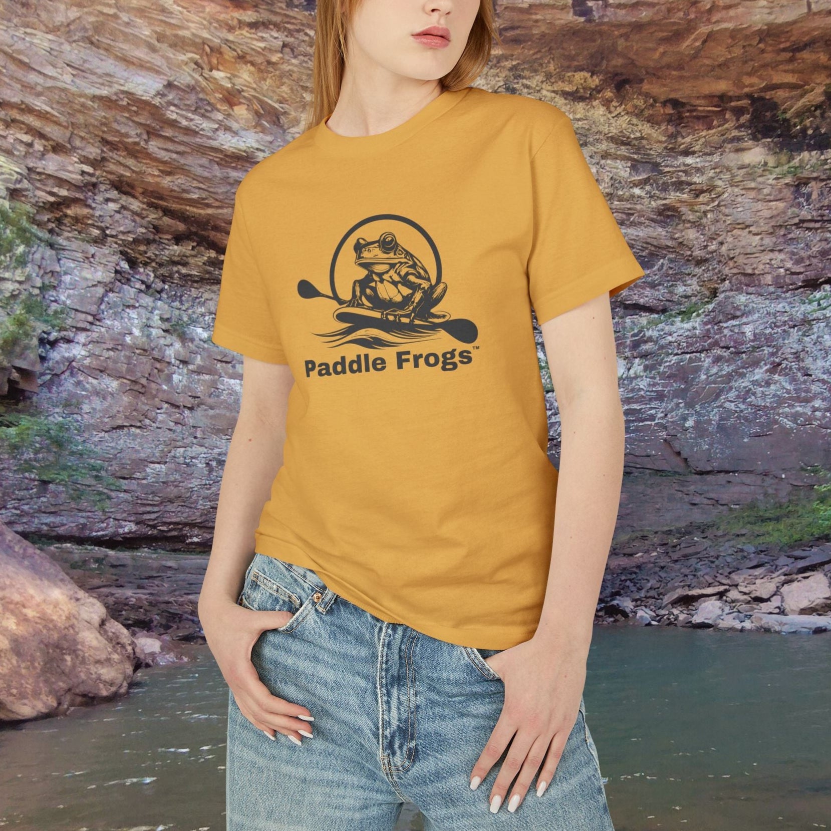 Woman wearing a yellow 'Paddle Frogs' t-shirt standing in front of a natural rock formation. Earthy Tee collection Faded mustard Yellow