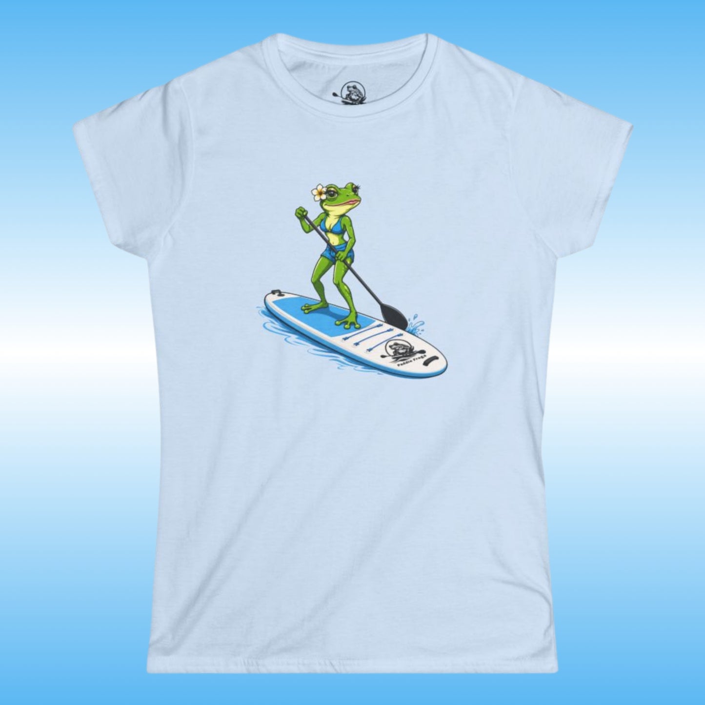 A light blue women's short-sleeve crewneck t-shirt featuring a cartoon graphic of a green frog paddleboarding on a blue and white board, set against a light blue gradient background.