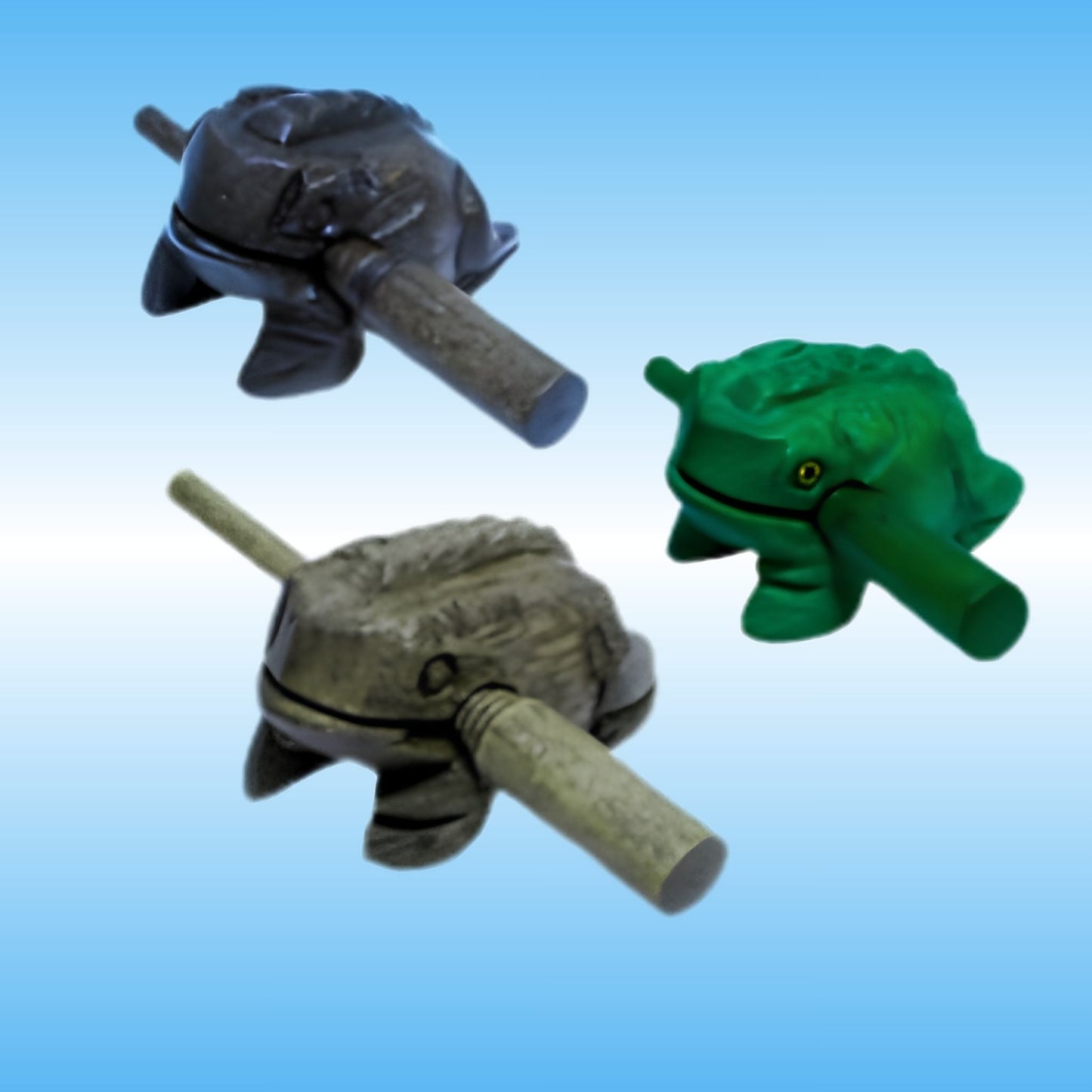 Three frog-shaped toy percussion instuments or novelty items shown on a light blue gradient background: one dark gray, one green with a small yellow eye detail, and one gray-green with a drum stick protruding from the front.