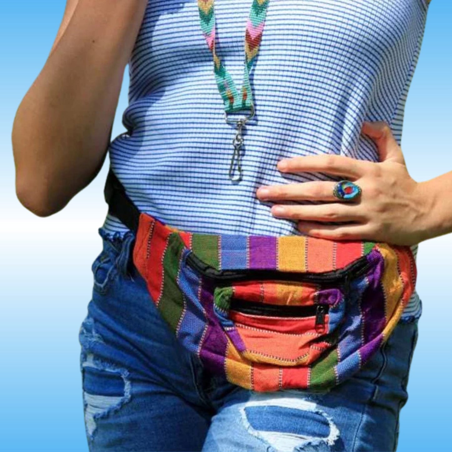 A close-up of a person wearing a colorful striped fabric waist bag pouch over blue jeans, with a blue-and-white striped top and a multicolor lanyard visible.