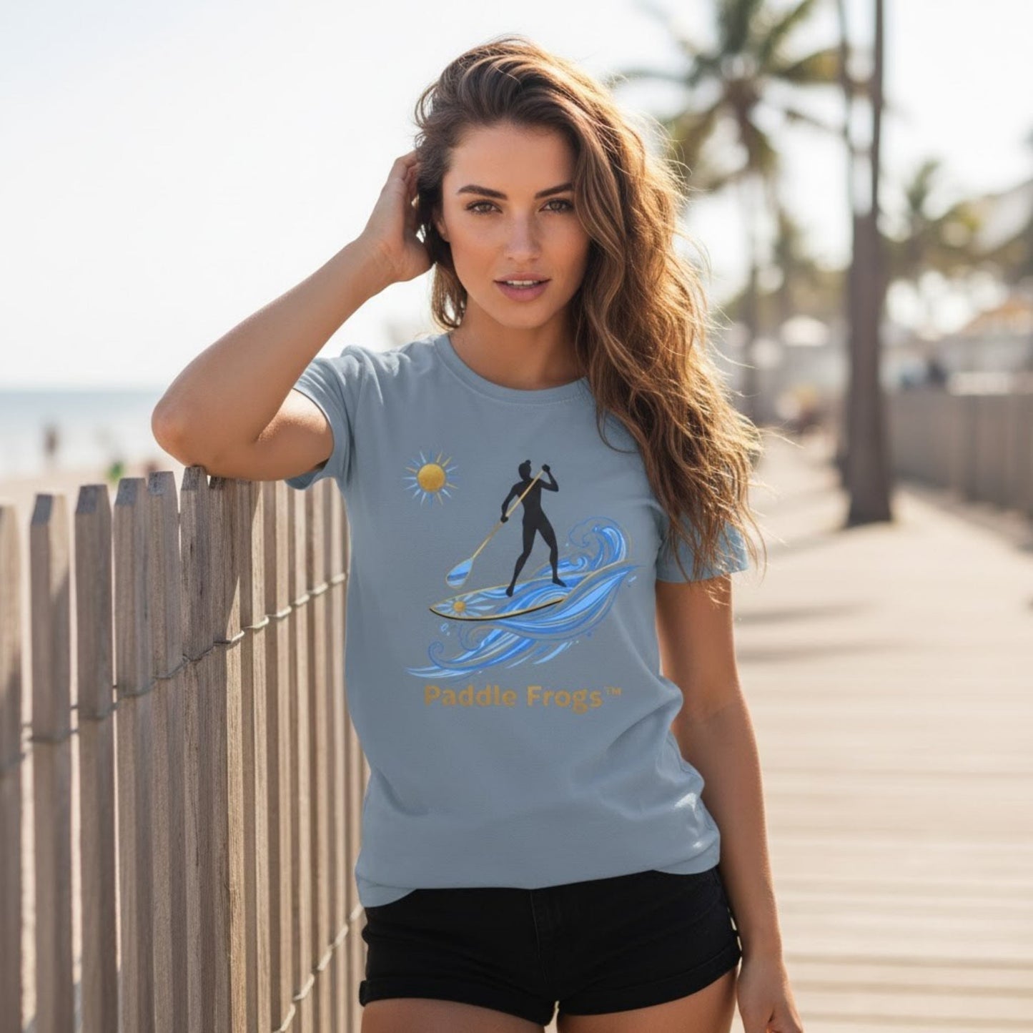 Woman wearing a light blue t-shirt with a graphic design on a beachside boardwalk. | at the beach "Sun Stoke" High-Quality Stand-Up Paddleboard T-Shirt – Premium Ring-Spun Cotton SUP Tee