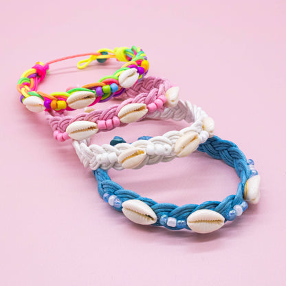 Four braided cord bracelets with cowrie shells, arranged in a fan shape on a solid pink background. The bracelets are in pink, white, blue, and a bright multicolor mix.