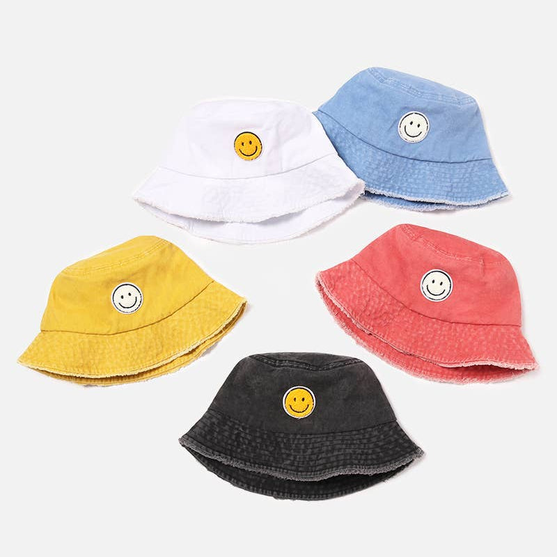 Five small bucket hats in white, light blue, yellow, coral pink, and dark charcoal gray, each featuring a small embroidered yellow smiley face on the front, arranged on a light gray background.