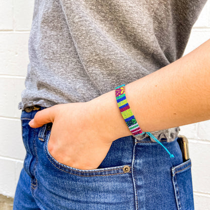 A close-up of a person's wrist wearing a colorful braided beaded bracelet, paired with a gray t-shirt and blue jeans against a white brick wall background.