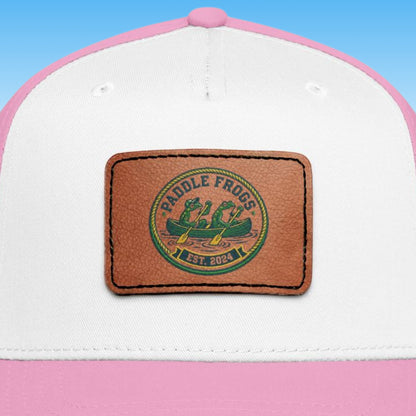 A close-up of a trucker-style cap with a white front panel, pink brim and top button, and a rectangular brown leather patch on the front. The patch has a circular green and yellow logo reading 'Paddle Frogs Est. 2024' with two frogs canoeing.
