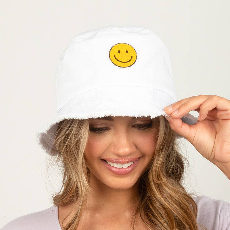 A woman wearing a white bucket hat with a yellow smiley face patch on the front, holding the brim with one hand. She is wearing a light lavender top against a plain light background.