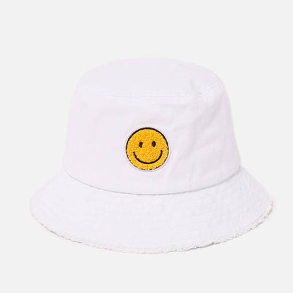 A white bucket hat with a yellow embroidered smiley face patch on the front, set against a light gray background.