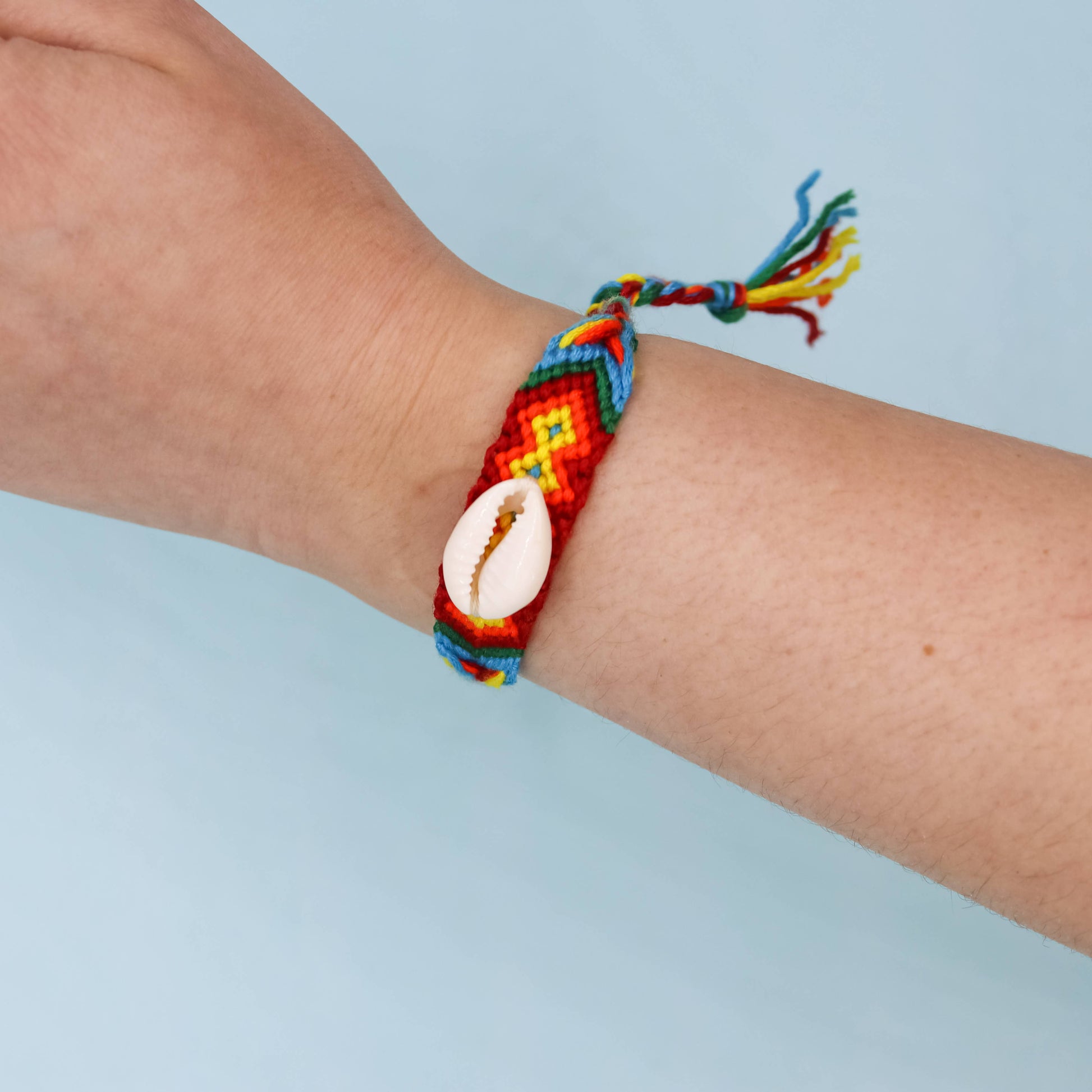 A multicolor braided friendship bracelet with a white cowrie shell centerpiece, worn on a person's wrist against a light blue background.