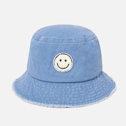A light blue denim bucket hat with a distressed frayed brim and a circular embroidered smiley face patch on the front, set against a plain light gray background.