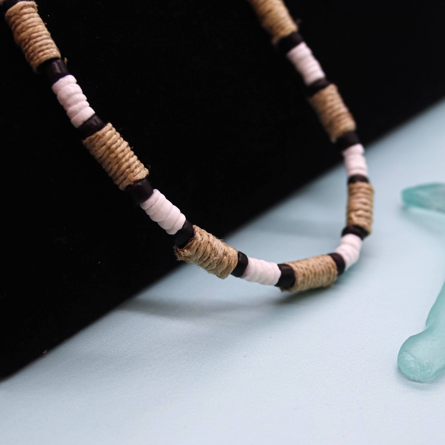 A close-up of a beaded necklace with alternating natural tan wrapped beads, white beads, and black spacer beads, displayed on a black surface against a pale blue background with a translucent turquoise object partially visible in the corner.