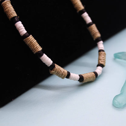 A close-up of a beaded necklace with alternating natural tan wrapped beads, white beads, and black spacer beads, displayed on a black surface against a pale blue background with a translucent turquoise object partially visible in the corner.