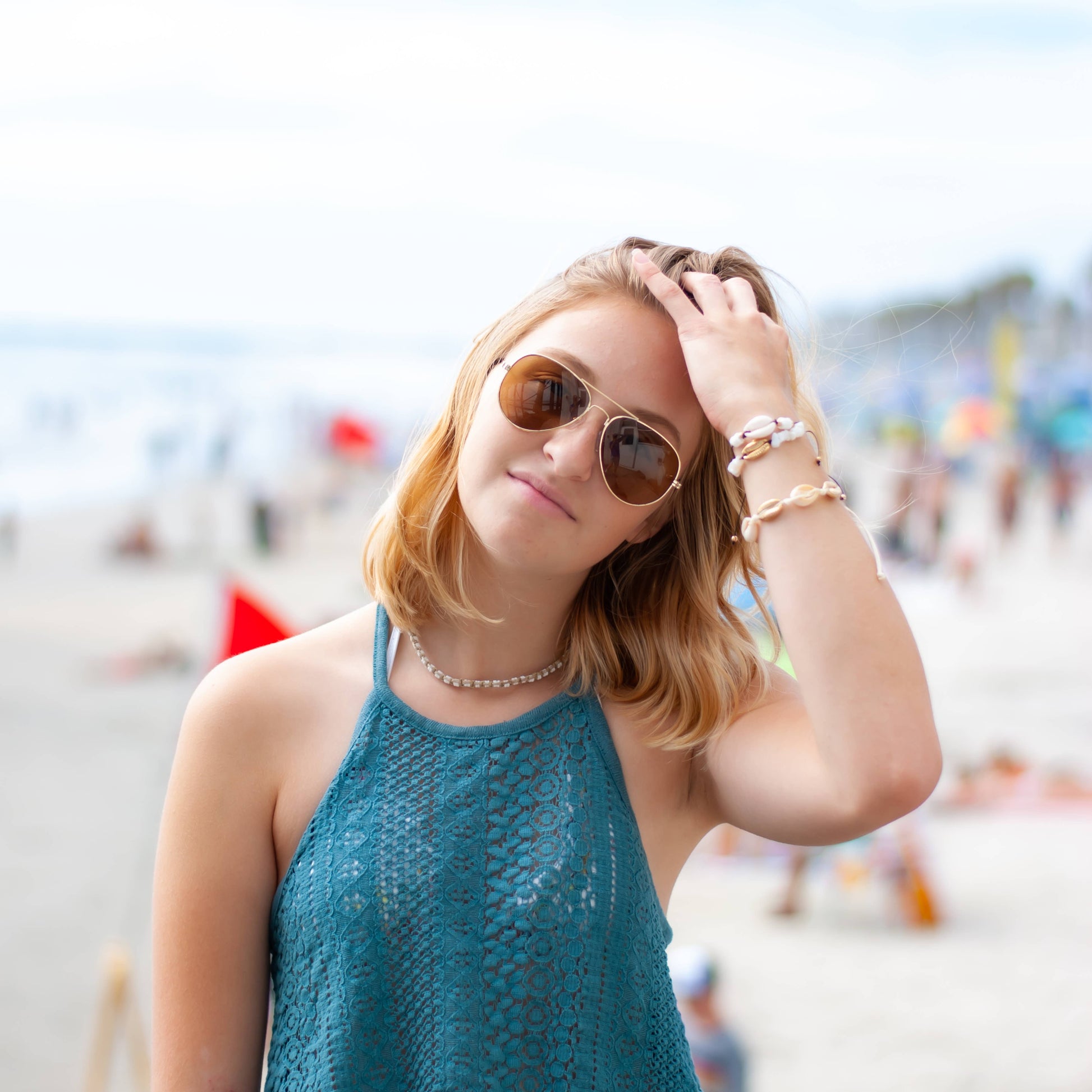 Woman wearing a hemp puka shell necklace, sunglasses and a blue top with a blurred beach background