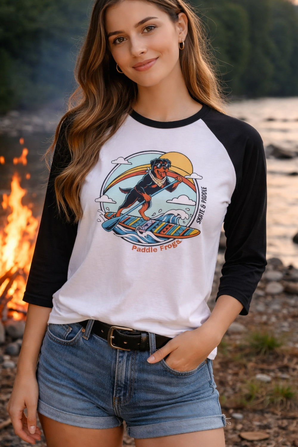 A young woman standing by a river and a warm campfire, wearing a white and black 3/4 sleeve raglan shirt with a graphic illustration of a Rottweiler dog stand-up paddleboarding on a wave. She is also wearing denim shorts and a black belt.