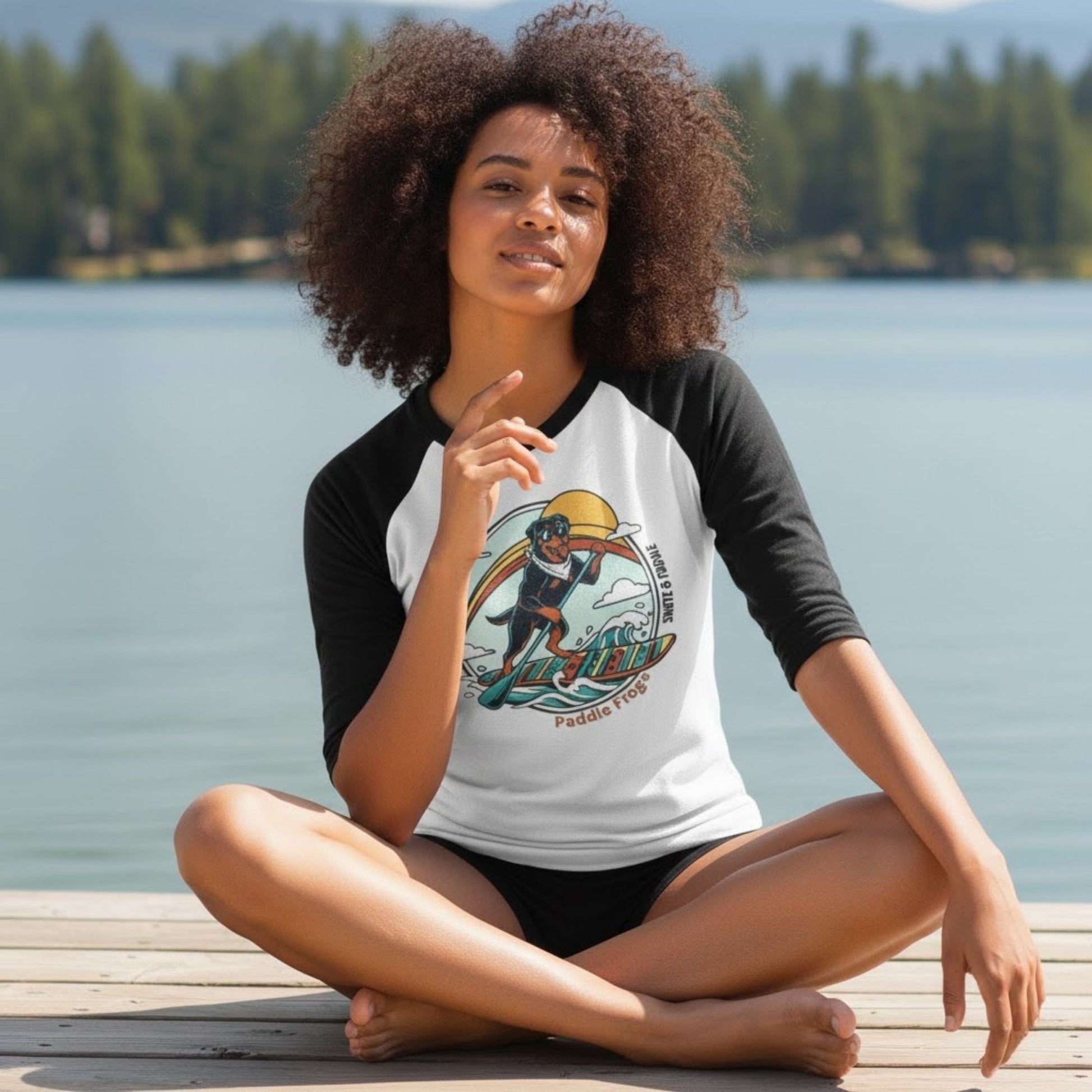 Woman sitting on a dock by a lake wearing a black and white baseball shirt with a graphic design of a rottweiler paddleboarding.
