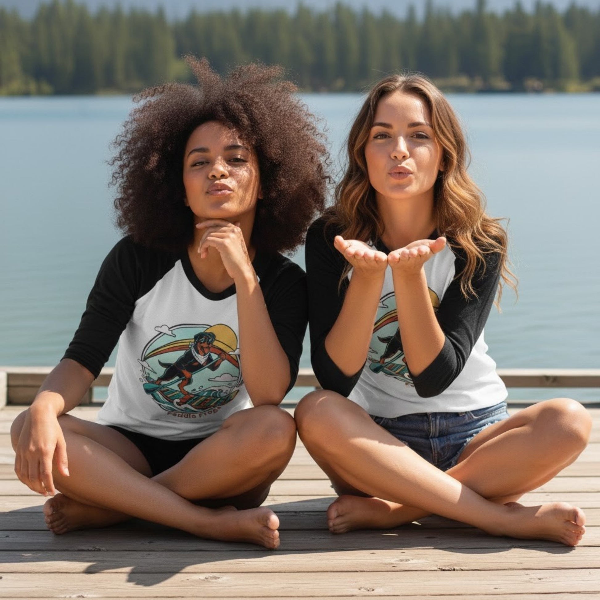 Two women sitting crisscross applesauce on a dock by a lake, wearing matching baseball jerseys.
