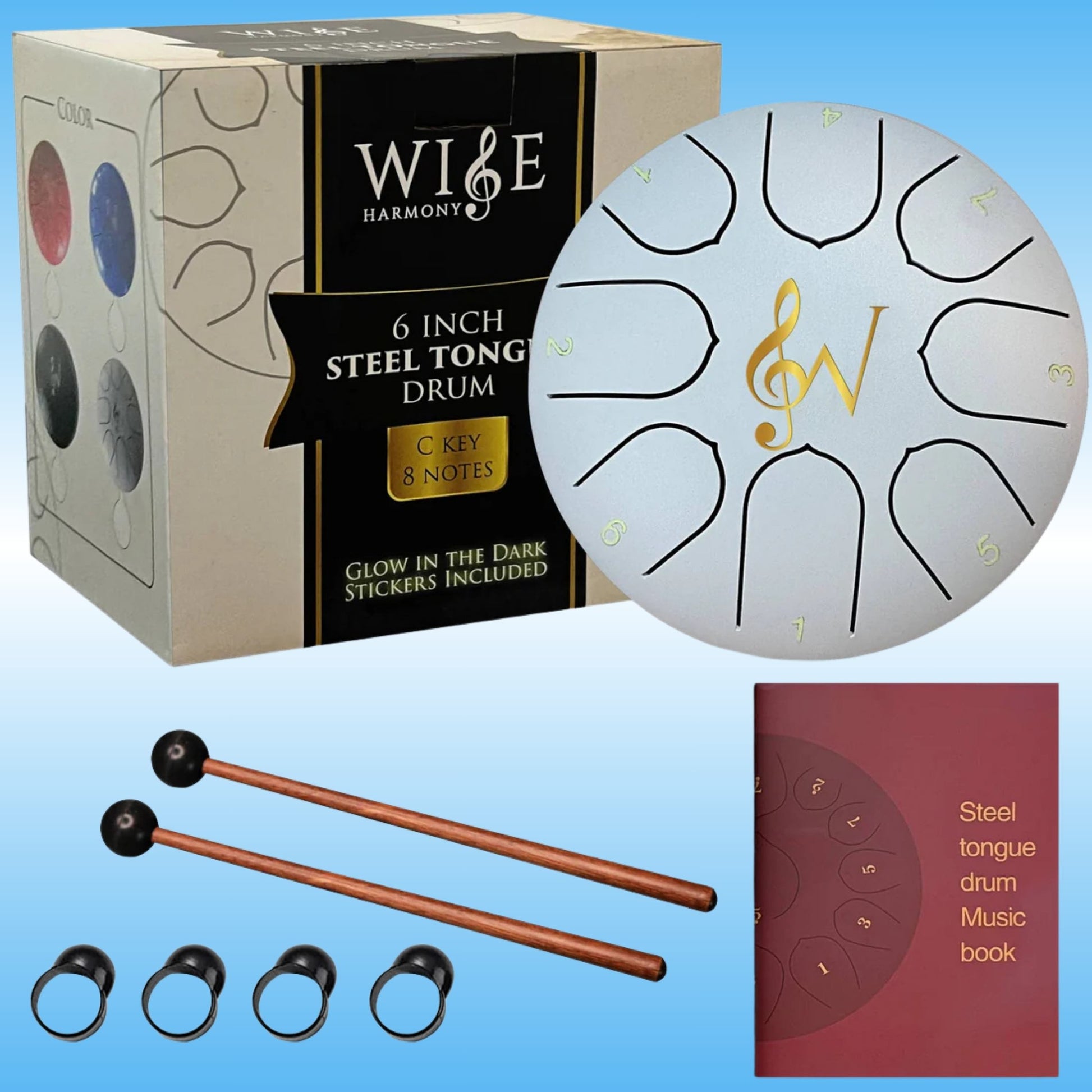 A steel tongue drum set with its packaging, shown on a light blue gradient background. The set includes a round white drum with black and gold markings, two copper-colored mallets with black tips, four black finger rings, and a red music book.