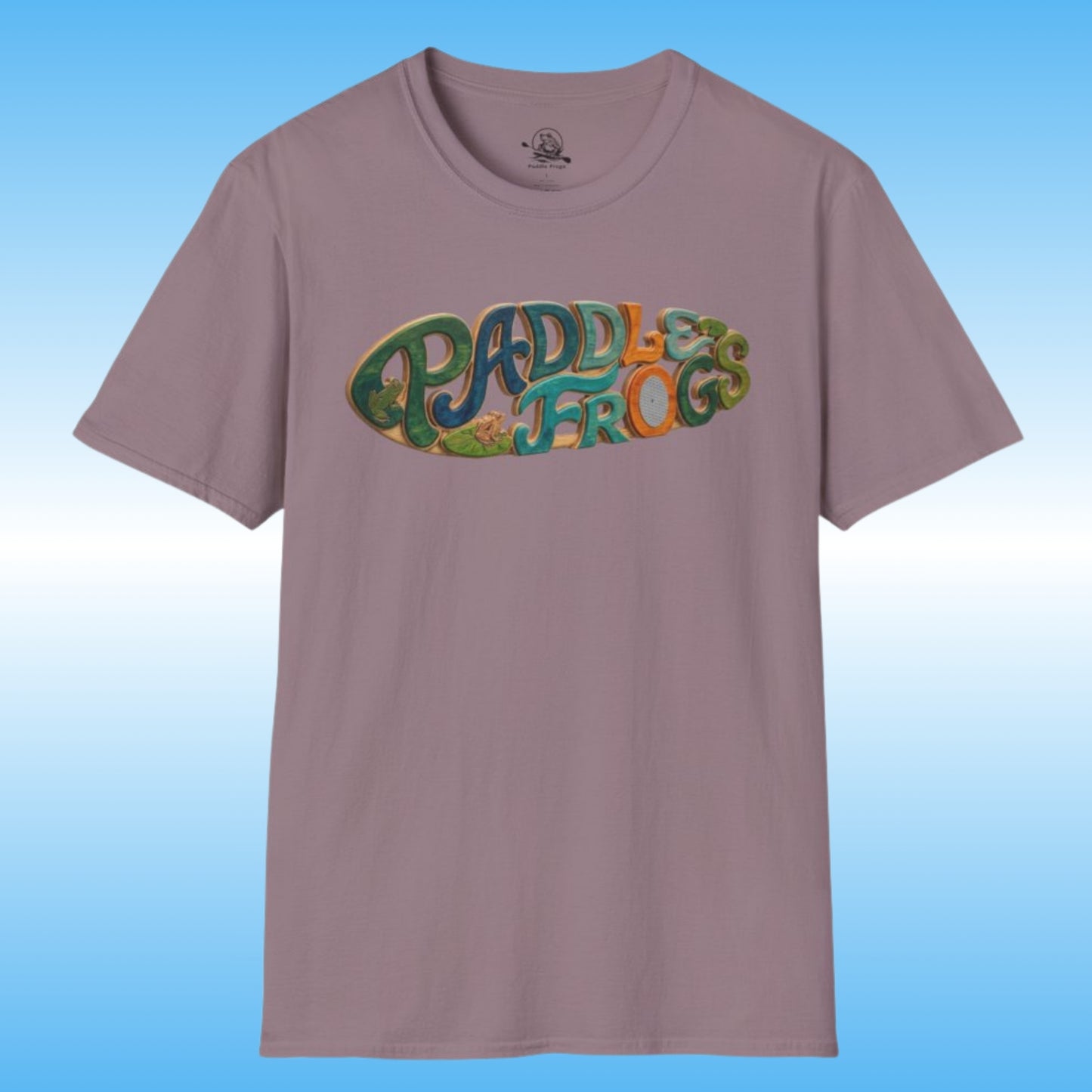 A short-sleeve crewneck t-shirt in a dusty mauve or muted purple color, featuring a colorful graphic on the chest that reads 'PADDLES FROGS' in a retro, bubbly font with green, blue, and orange details creating a surf paddleboard. The shirt is displayed against a light blue gradient background.