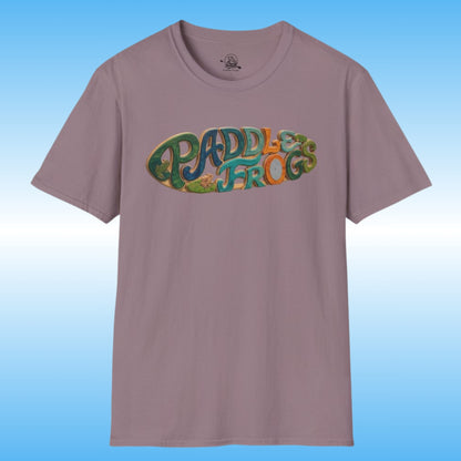 A short-sleeve crewneck t-shirt in a dusty mauve or muted purple color, featuring a colorful graphic on the chest that reads 'PADDLES FROGS' in a retro, bubbly font with green, blue, and orange details creating a surf paddleboard. The shirt is displayed against a light blue gradient background.