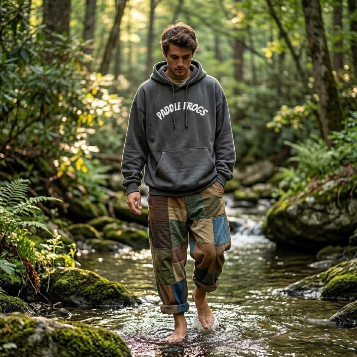 A man walking barefoot in a forest stream wearing a dark smoke gray hoodie with 'Paddle Frogs' printed on the chest and patchwork camouflage pants.