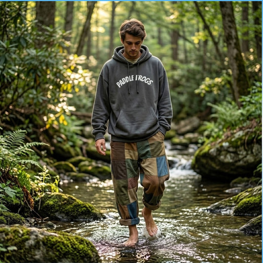 A man walking barefoot in a forest stream wearing a dark smoke gray hoodie with 'Paddle Frogs' printed on the chest and patchwork camouflage pants.